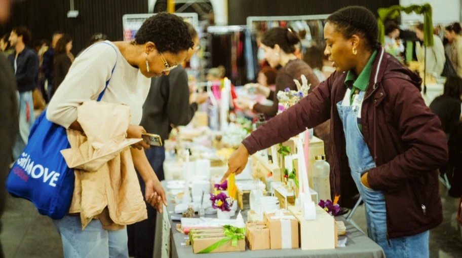 Vendedora a mostrar velas e presentes a uma visitante, com o mercado cheio ao fundo